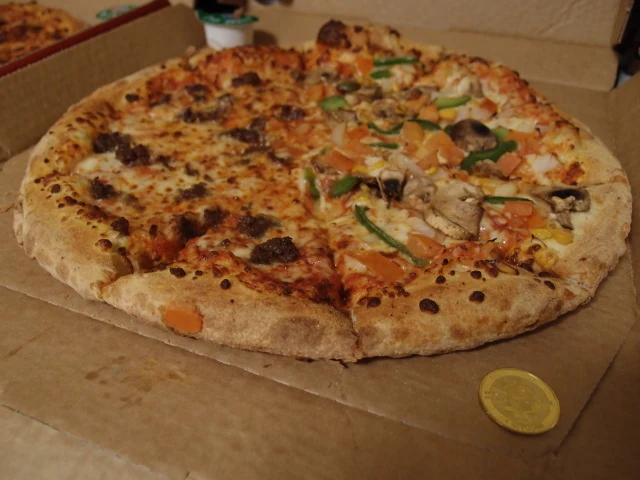 A half-and-half pizza in its delivery box: ground beef on one side, veggie supreme with peppers, mushrooms, onion and tomato on the other, with a gold Casascius bitcoin beside it